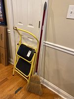 Yellow metal two-step ladder leaning against a door with black non-slip steps. Next to it is a traditional broom with natural fiber bristles and red handle, leaning against the wall.