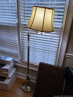 Full view of brass standing lamp with fabric shade beside window.