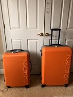 Front view of two orange Nautica hard shell suitcases with telescoping handles extended on a beige carpet in front of white double doors.