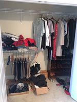 Wide view of closet showing hanging suit jackets, shirts, folded clothing, two boxes on floor with various clothes, and shoes on bottom shelf and floor.