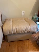 View of beige leather ottoman showing top cushion and base on wood floor