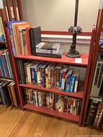 Photo of a red wooden bookshelf holding various books on all three shelves and a decorative lamp on the top shelf.