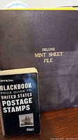 Large black Deluxe Mint Sheet File with gold text on front cover, along with a 2007 Official Blackbook Price Guide to US Postage Stamps booklet visible below.