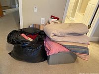 Photo showing black bag filled with various towels, a plastic bin with folded pink sheet and striped blanket on top with beige towels on the side.
