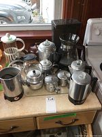 Overview photo showing multiple stainless steel and glass coffee and tea items arranged on a kitchen countertop in front of a window with a stove to the right.