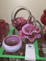 Photo showing cranberry glass baskets with handles and hobnail pattern bowl with pink and white opalescent finish, displayed on glass shelf.