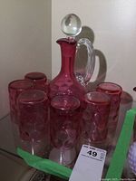 Cranberry glass decanter with clear stopper and handle, surrounded by six matching cranberry glasses, viewed from front
