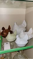 Five hen on nest dishes on glass shelf including four white and one brown ceramic/pottery.