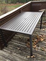 Full view of the rectangular metal patio table on a weathered wooden deck, showing the slatted top surface and umbrella hole.