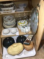 Wide view of cupboard interior showing stacked plates, egg cups, a white ceramic double section dish, serving domes, lidded bakeware and wooden bowls.