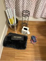 Photo shows metal bucket with two rolls of white and gray patterned contact paper, black metal tissue holder, metal toilet paper holder, and black Steve Madden plush rug partially visible.