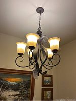 Full view of a 5-light chandelier hanging from ceiling with amber colored shades and wrought iron frame.
