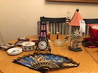 Image showing tabletop clock in black metal frame styled as easel, textured fan, and decorative birdhouse with barometer along with other decorative items on the table.