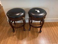 Front view of two wooden counter stools with black round cushioned tops and carved leg details on a hardwood floor.