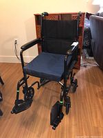Front view of the black folding wheelchair with armrests and footrests, showing attached blue gel seat pad.