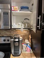 Photo of kitchen counter with Keurig coffee maker and collection of mugs and cups beneath shelf.