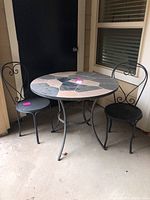 Photo showing the round stone tiled tabletop with black metal frame and two black metal patio chairs with decorative backs.