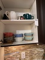 Two shelves showing white, blue, and dark ceramic teapots, a floral cup on the top shelf, and multiple stacked soup bowls in red, blue, gray, and green tones on the lower shelf.