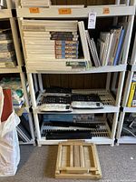 Shelving unit with stacks of white stretched canvases and canvas boards on top shelf, spiral-bound pads on right, brush sets on second shelf and wooden travel easel on floor.