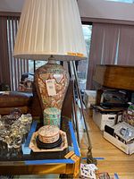 Asian-style lamp with detailed ceramic base and pleated lampshade, shown beside other ceramic decorative items on a glass table.