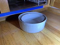Photo showing two white round casseroles nested together with visible vertical ridges on the outer one at base corner in wood flooring setting.