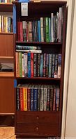 Full view of dark wood bookcase with three shelves and two drawers, filled with three rows of books mostly hardbacks.