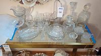 Wide view of assorted clear glass and crystal items on a glass table including decanters, bowls, candy dishes, and serving trays.