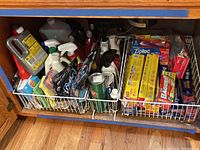 View of assortment of cleaning supplies and food wrap boxes inside a lower kitchen cabinet on two white wire baskets.