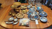 Full view of assorted metal serving pieces including bowls, trays, coffee pots, and a plastic bag with forks on a wooden table.