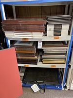Shelving rack with stacked frames on top and middle shelves alongside portfolios on bottom shelf. Frames are boxed or shrink-wrapped with visible wood finishes in brown and light colors. Black portfolios in bottom shelf.