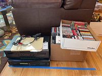 Photo of Sony stereo cassette deck TCWR521, Sony CD/DVD player DVP-NC665P, and large box of assorted CDs and tapes sitting on floor in front of chair.