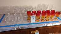 Wide view of all stemware arranged on table including clear wine glasses, brandy snifters, champagne flutes, and colorful orange-yellow short-stemmed glasses.