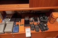 Wide view of all vintage transistor radios and binoculars on wood shelf.