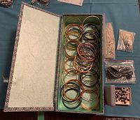 Fabric-covered box open with over 50 assorted metal and ceramic bangles arranged inside, clear view of various colors and styles, plastic bags containing white and dark beads alongside the box, and a small jewelry storage tray with pearl-like beads on the side.