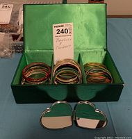 Green fabric jewellery box open with over 30 colorful bangles arranged inside in compartments, also showing a heart-shaped compact mirror in front.