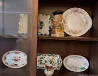 Two shelves displaying assorted Serving dishes, trays and a Toby mug. Floral patterns dominate, some dishes are oval, others rectangular, featuring gold trimmed edges.