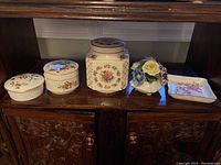 All five items displayed on a wooden shelf: two covered jars, one covered dish, a flower figurine, and a small plate all with floral designs.