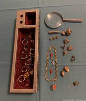 Overview of the metal box containing rings and assortment of jewelry items including bracelet, earrings, pins, and magnifying glass laid out next to the box.