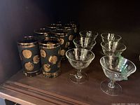 Photo showing 8 vintage black tumblers with gold circular designs and gold rims alongside 6 clear crystal dessert coupes with etched cross and olive designs