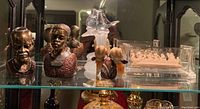 Various figurines on a glass shelf including two metal busts, glass birds on stone base, and acrylic encased boat figurine