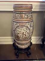 Front view of tall ceramic vase with intricate hand-painted floral and bird designs on white background, mounted on carved wooden stand.