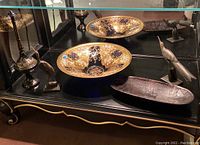 Wide shot of all items in a display case: cobalt blue glass dish, two metal bird figurines, and wood tray on black surface with mirror background