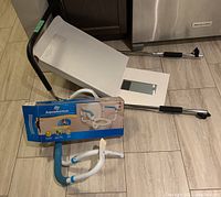 Shows metal ramp with black handles and support legs, the boxed toilet seat, and the white box containing the toilet lift or other device, placed on kitchen floor