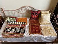 Shows embroidered pillow covers in cream and gold floral patterns and a black/red decorative pillow, along with multicolored woven throw blanket on a metal frame daybed.