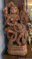 Front and angled views of the carved wooden Lakshmi sculpture showing detailed jewelry, shield, and ornamental patterns.
