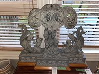 Front view of sculpture lamp, light off, showing carved figures and floral motifs on stone atop wood base