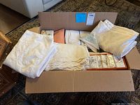 Full open cardboard box showing mixed folded white and cream-colored table linens including runners, tablecloths, and napkins, some still in packaging.