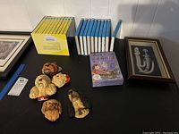 View of boxed Nancy Drew books with yellow spines, boxed Hardy Boys books with blue spines, Amelia Bedelia book deck, six ceramic dog head plaques, and framed dagger on tabletop against white wall.