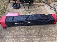 Full length view of black and red wheeled storage bag labeled ABCCANOPY on concrete surface outdoors.