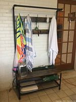 Full view of the hall tree against a white brick wall, showing towels hanging on hooks, wooden shelves, and metal frame base with wire mesh shelves.